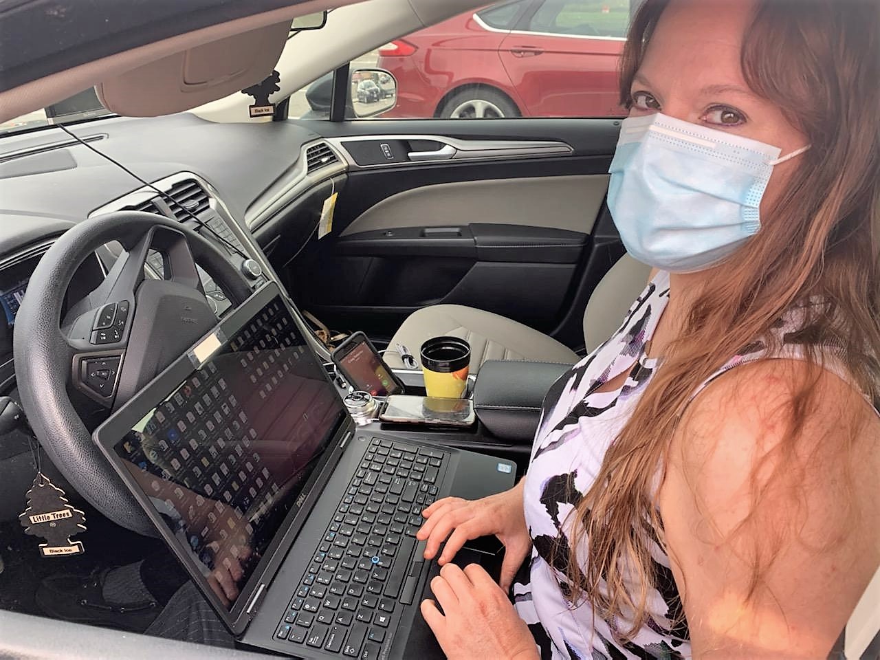 Woman sits in a car wearing a mask working on a laptop