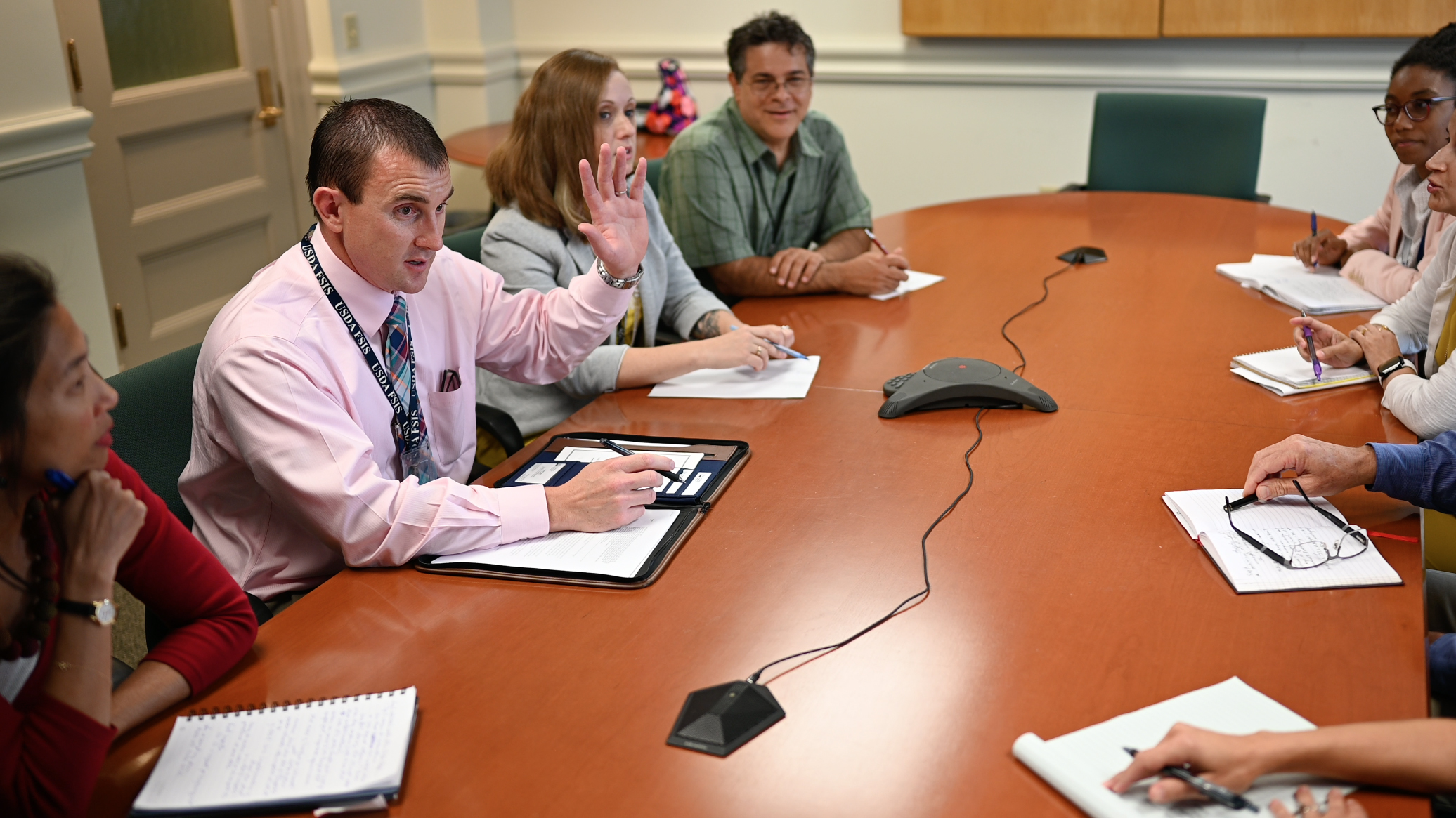 A group of people sits around a conference room table. One man has his hand raised.