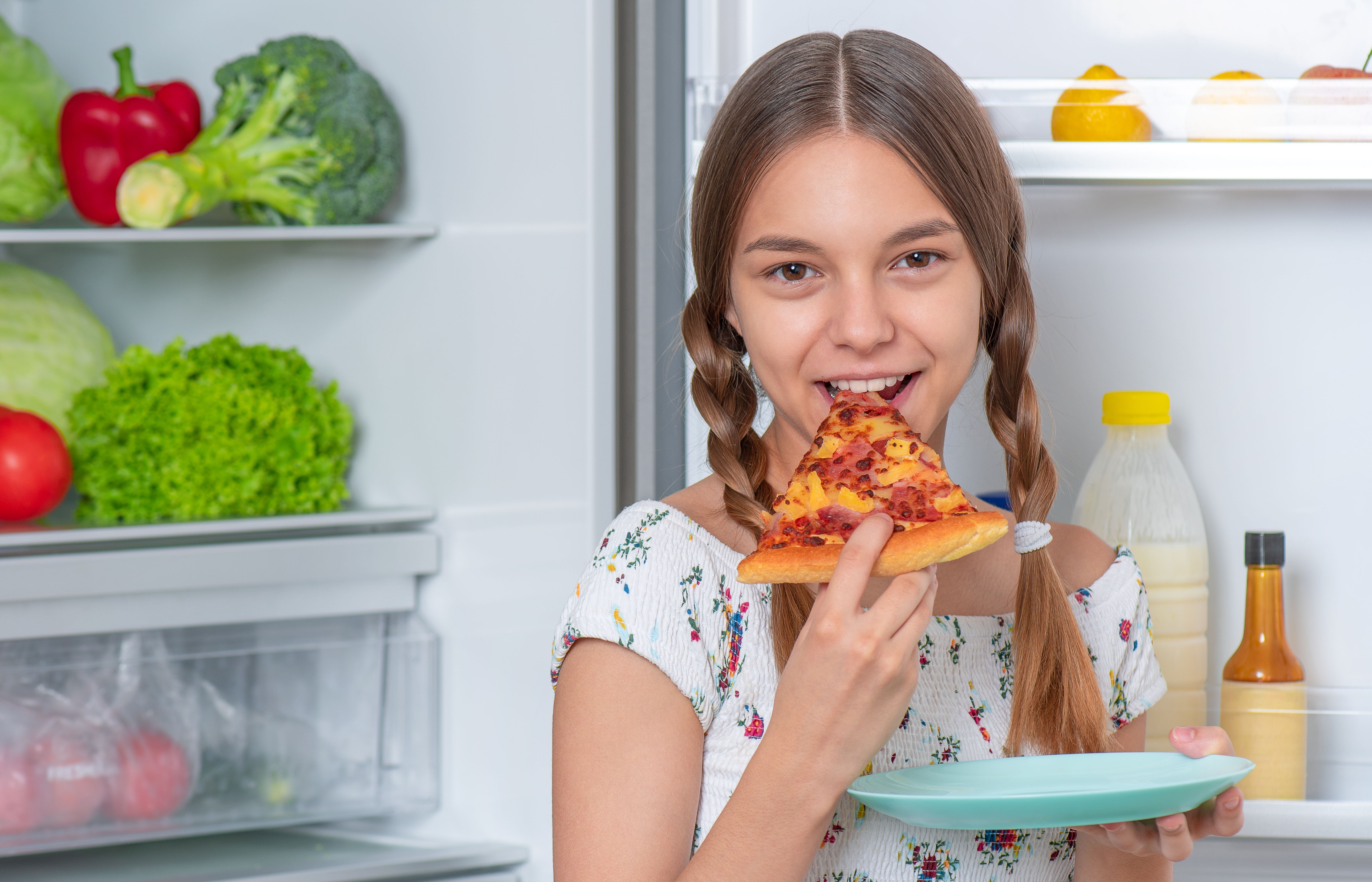 Teen girl eating pizza in front of a refrigerator